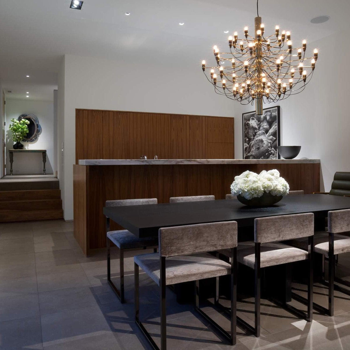 Warm bulbs light this gold-toned Clérachic chandelier over a dark dining table in a room with a walnut wall and a white flower bowl.