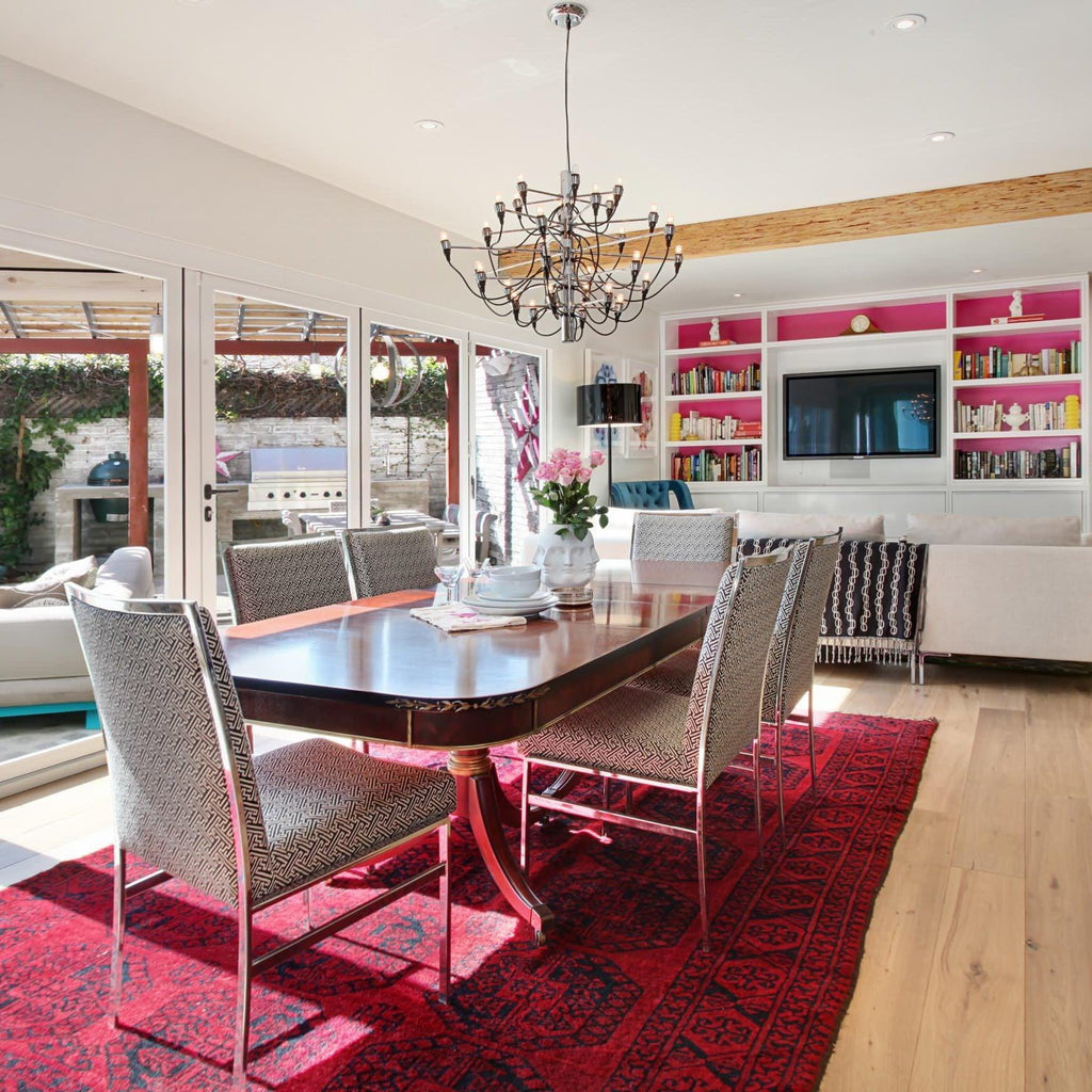 A Clérachic chandelier hangs over a dark wood dining table on a red rug, with patterned chairs and a pink-backed bookcase behind.