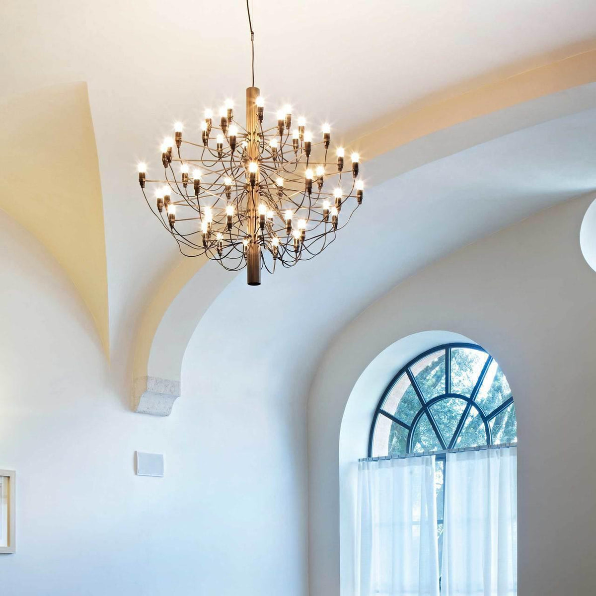 A bronze Clérachic chandelier with warm exposed bulbs hangs from a curved white ceiling, beside an arched window.
