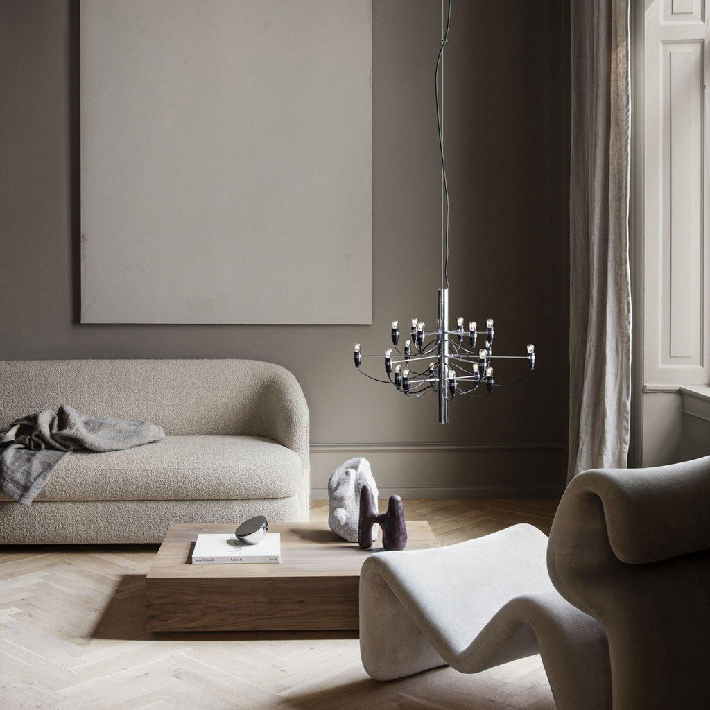 A slim Clérachic chandelier with a chrome stem hangs in a taupe living room, above a low wooden table and curved sofas.