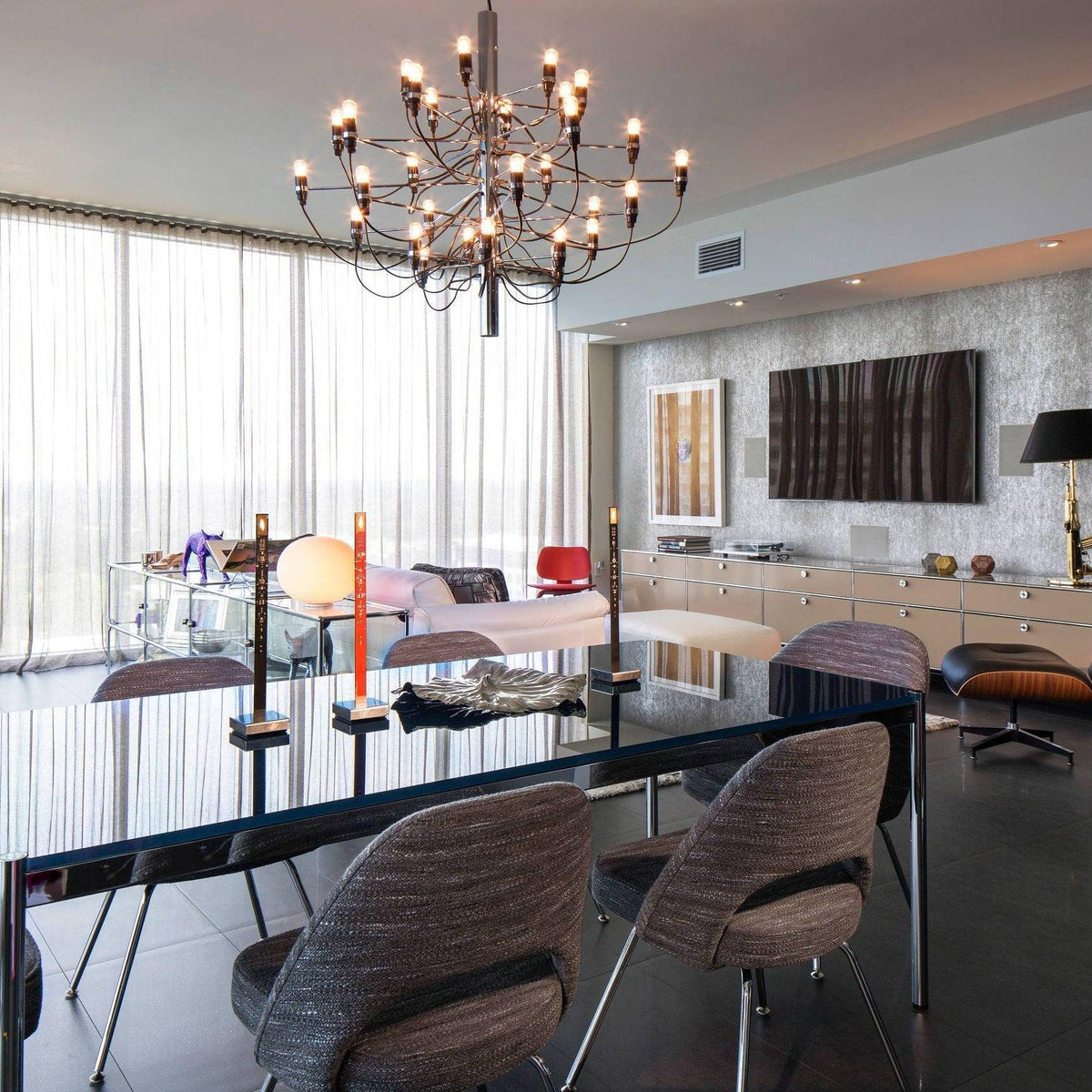 This Clérachic chandelier hangs in a bright room with floor-to-ceiling sheer curtains, above a dark glossy table and woven chairs.