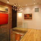 A lit Clérachic chandelier hangs near a glass balustrade, beside an exposed brick wall and a red painting.