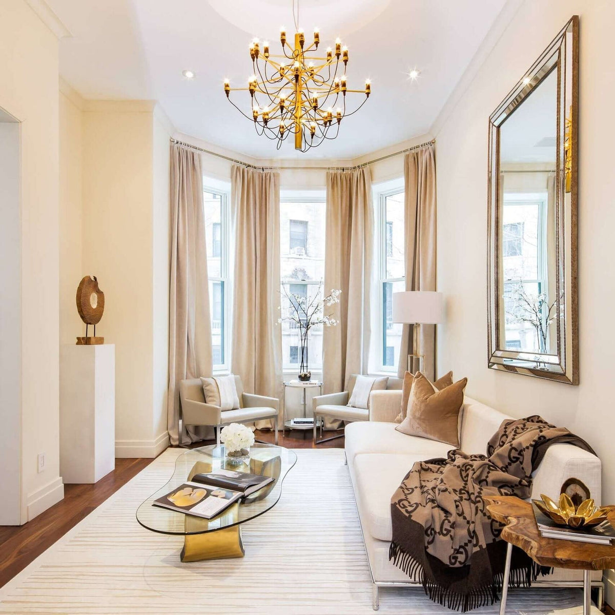 A gold Clérachic chandelier hangs high in a cream living room, above a glass coffee table, a white sofa and a large mirror.
