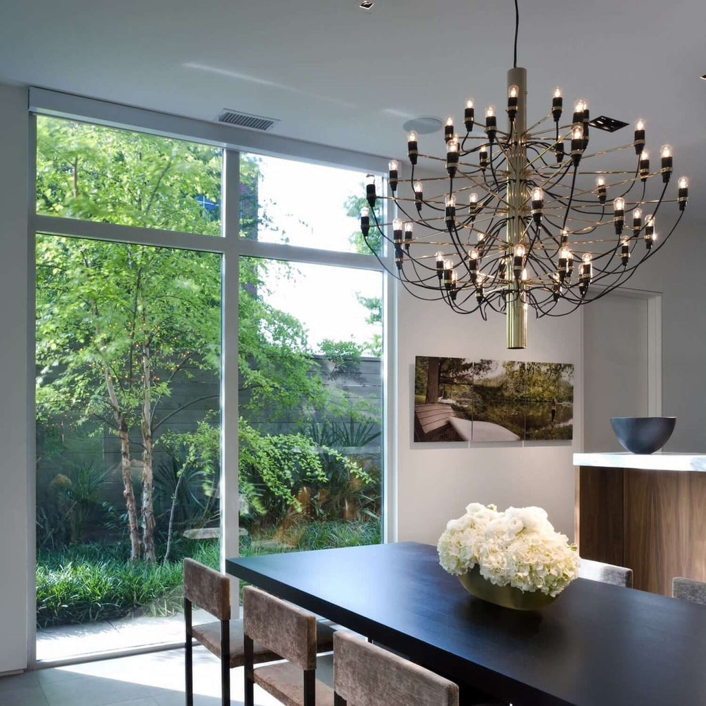 A Clérachic chandelier with a brass-toned stem hangs beside a garden window, above a black dining table with white flowers.