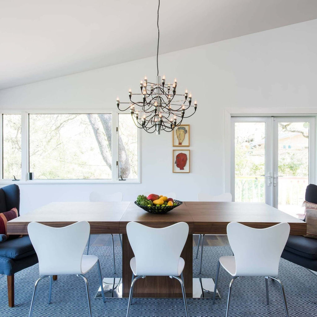 A Clérachic chandelier hangs from a sloped white ceiling over a walnut table with white chairs and a bowl of fruit.