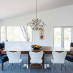 A Clérachic chandelier hangs from a sloped white ceiling over a walnut table with white chairs and a bowl of fruit.