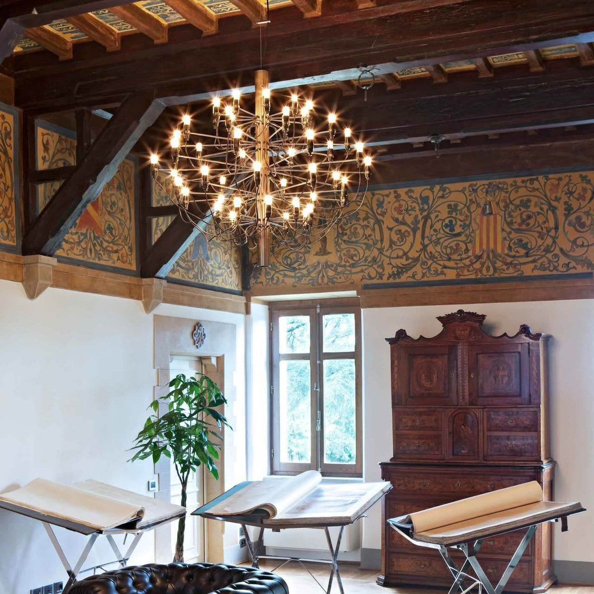 A Clérachic chandelier glows with warm bulbs under carved timber beams, in a room with painted walls and a cabinet.