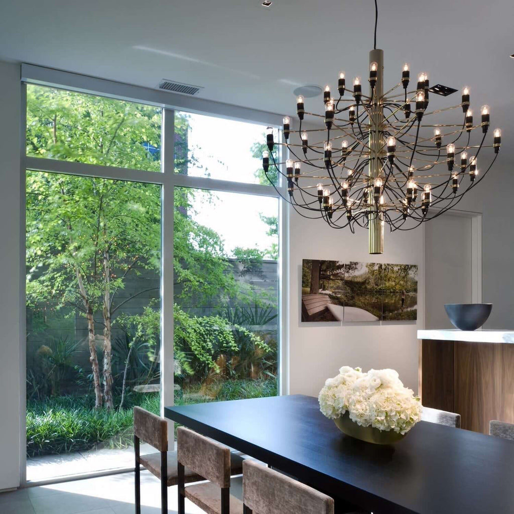 A Clérachic chandelier with a brass-toned stem, lit with warm bulbs, hangs beside a garden window and a dark table.