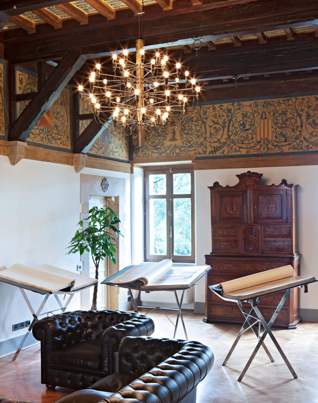 Seen from a wider angle, this Clérachic chandelier hangs in a timber-beamed room with leather sofas and wooden easels.