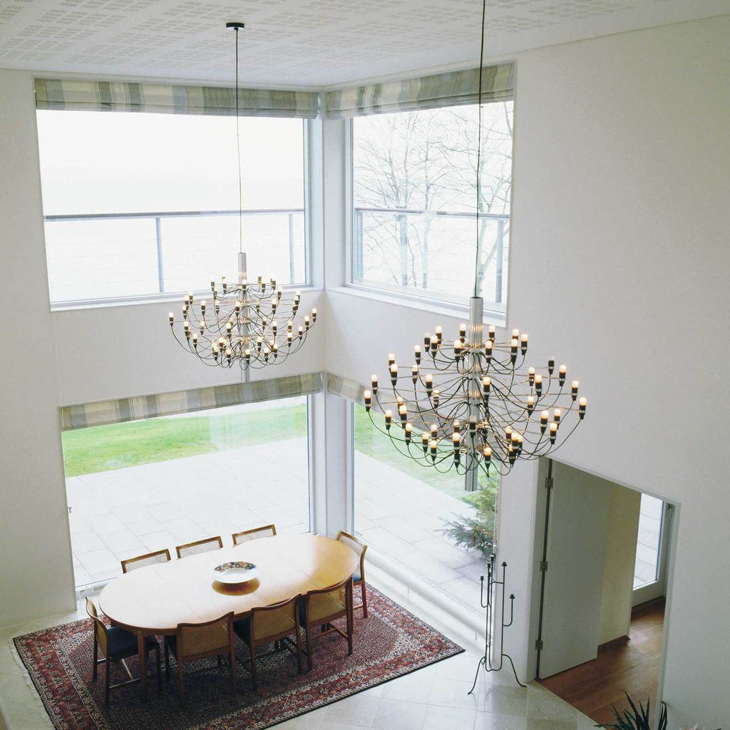 Two Clérachic chandeliers hang in a high-ceilinged room with corner windows, above an oval table on a patterned rug.