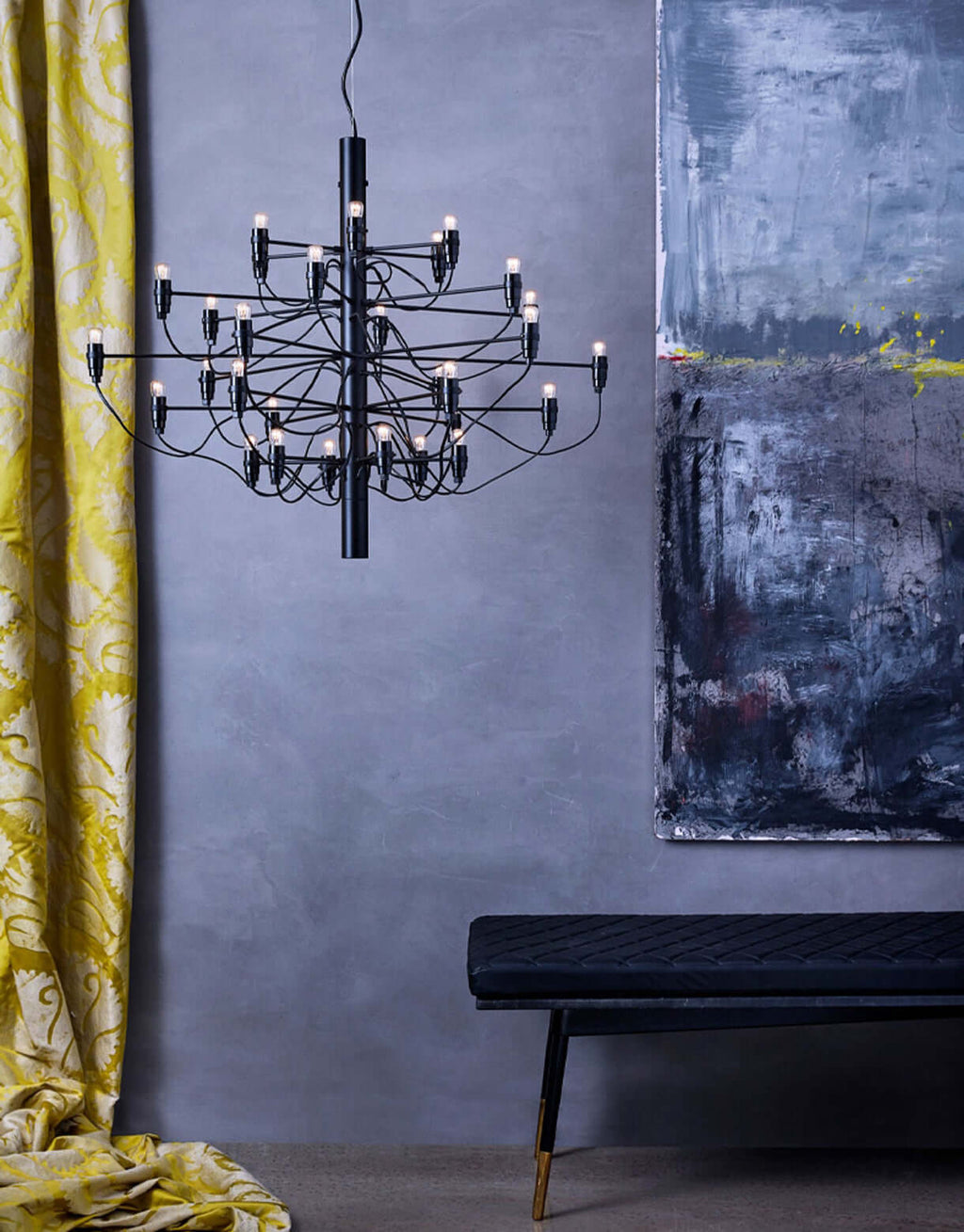 A matt black Clérachic chandelier hangs against a blue-grey wall beside a large abstract painting and a black bench.