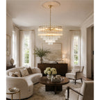 A Clérachic tiered glass chandelier lit warm above a round dark coffee table in a cream living room with a curved sofa.