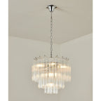 Chrome-chained Clérachic chandelier with tiers of clear glass rods, lit, hanging in the corner of a white room.