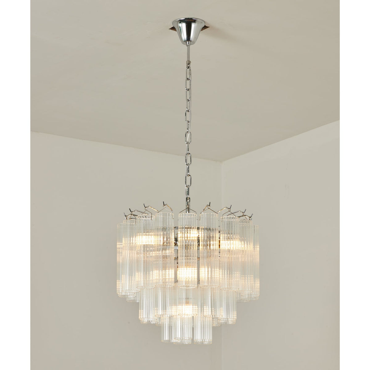 Chrome-chained Clérachic chandelier with tiers of clear glass rods, lit, hanging in the corner of a white room.