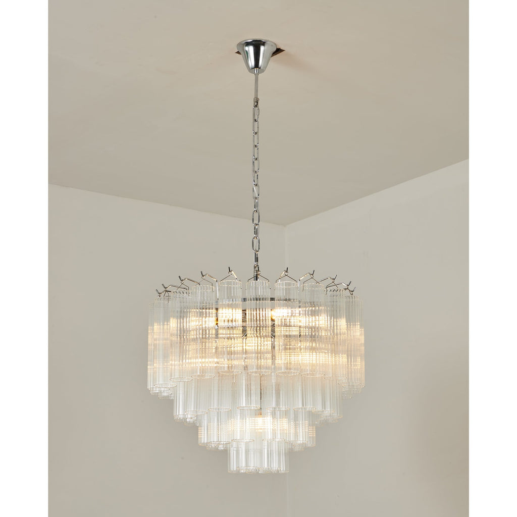 A Clérachic chandelier with chrome fittings and layered clear glass tubes, glowing warm against a cream ceiling corner.