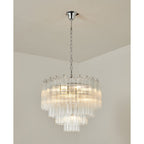 A Clérachic chandelier with chrome fittings and layered clear glass tubes, glowing warm against a cream ceiling corner.