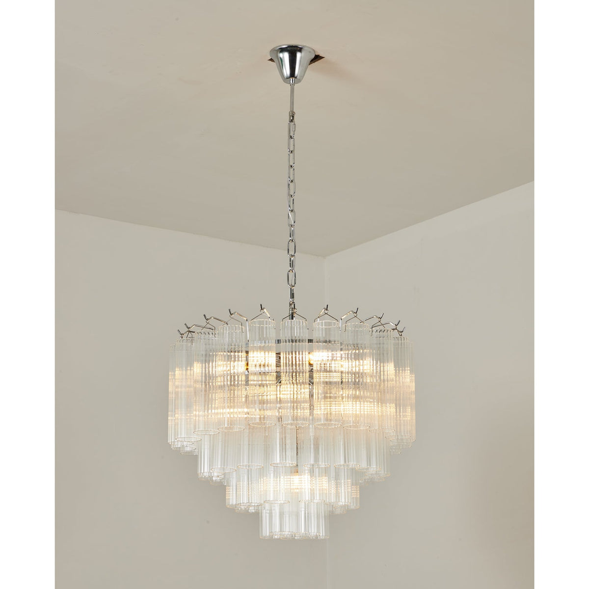 A Clérachic chandelier with chrome fittings and layered clear glass tubes, glowing warm against a cream ceiling corner.