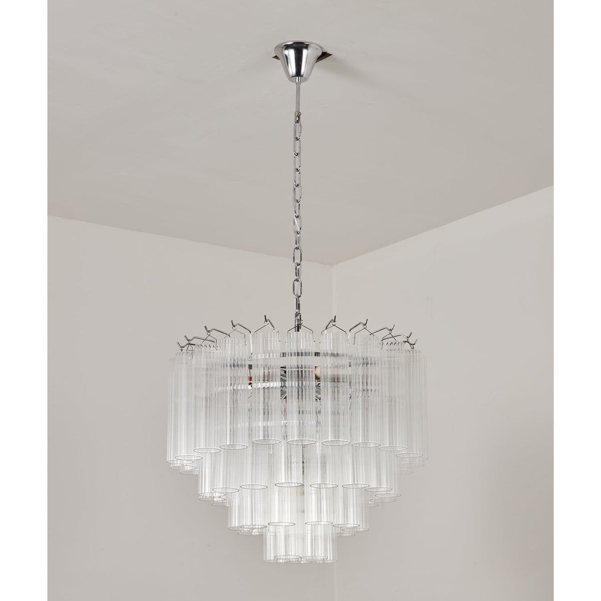 Unlit tiered glass chandelier from Clérachic on a chrome chain, the clear rods catching soft light in a plain room.