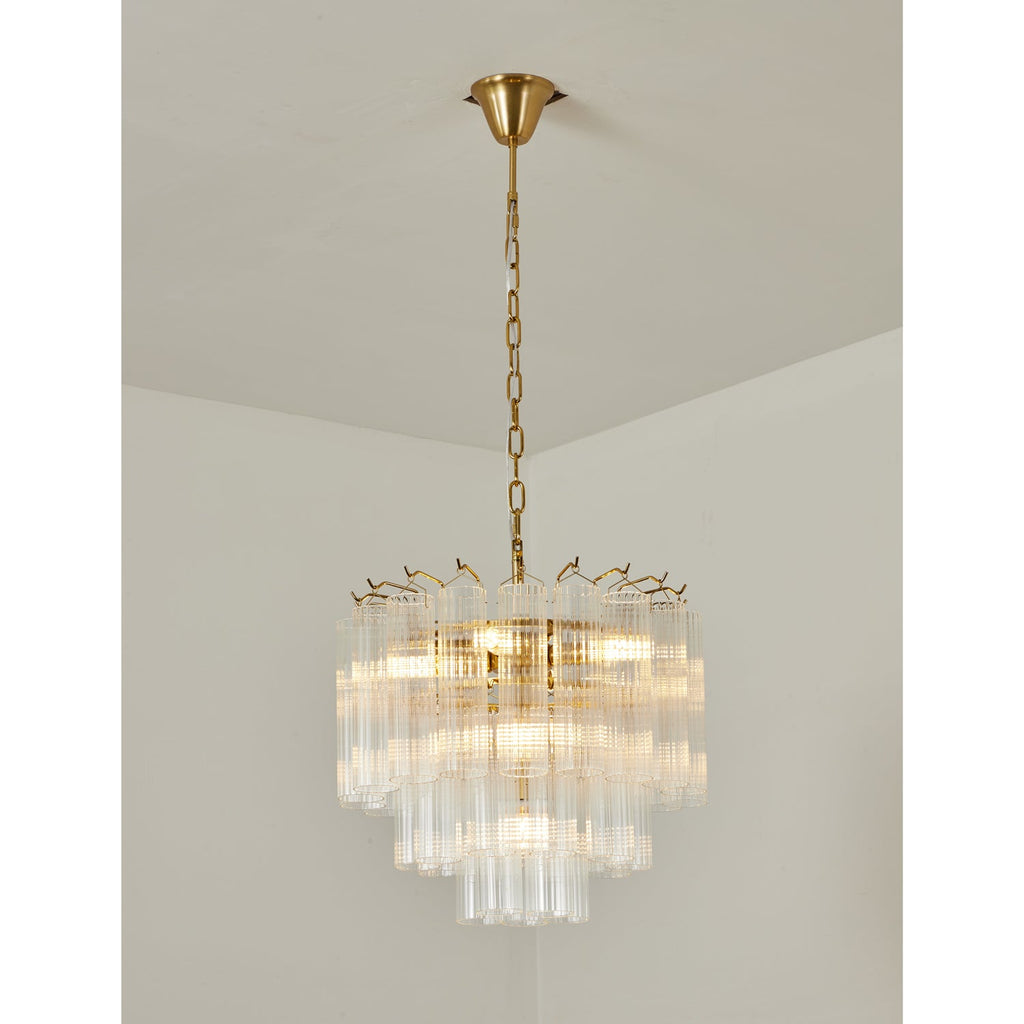 Warmly lit Clérachic chandelier with a gold-toned frame, its tiers of clear fluted glass glowing against a cream wall.