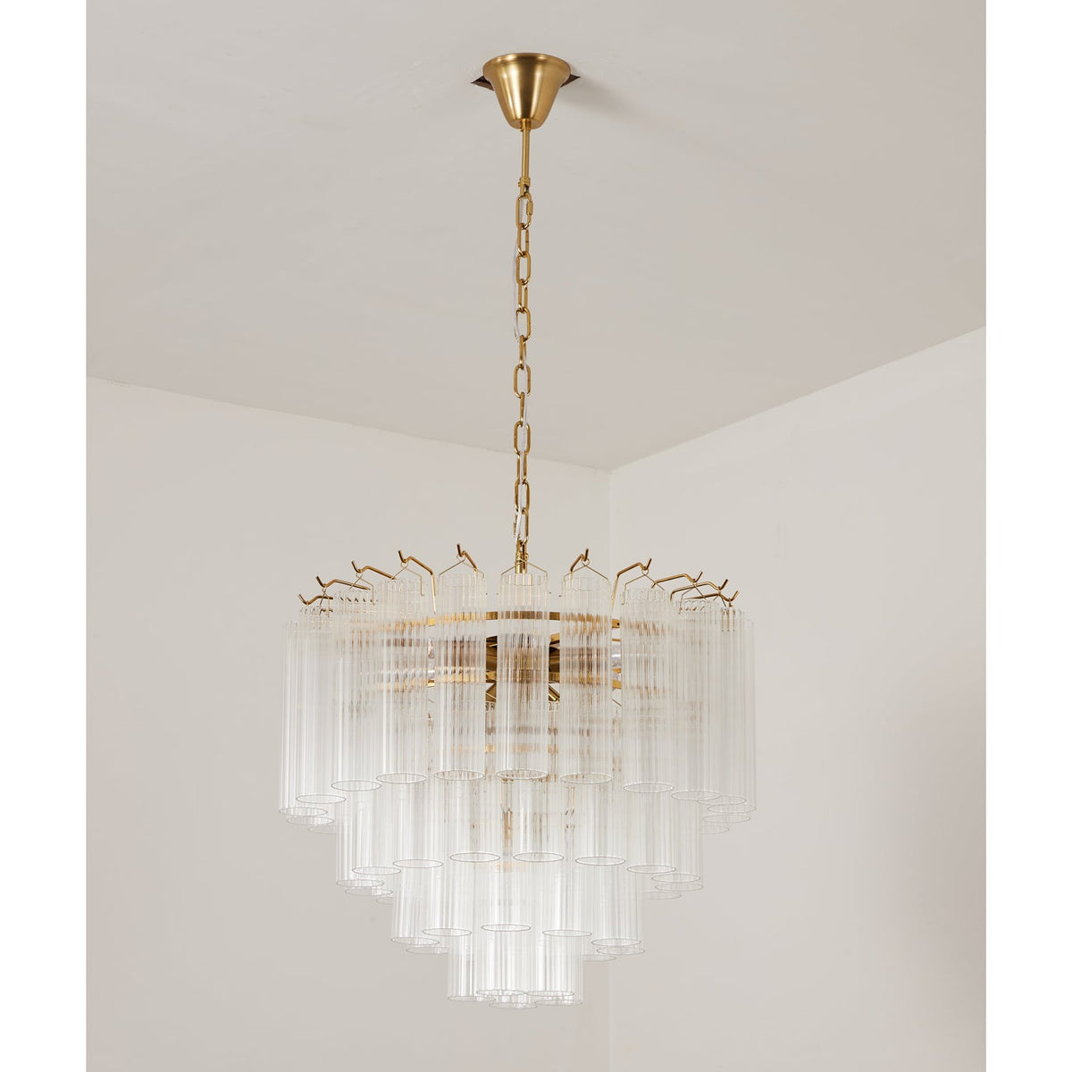 Compact unlit Clérachic chandelier with clear glass rods and a gold-toned chain, shown in a bare pale corner.