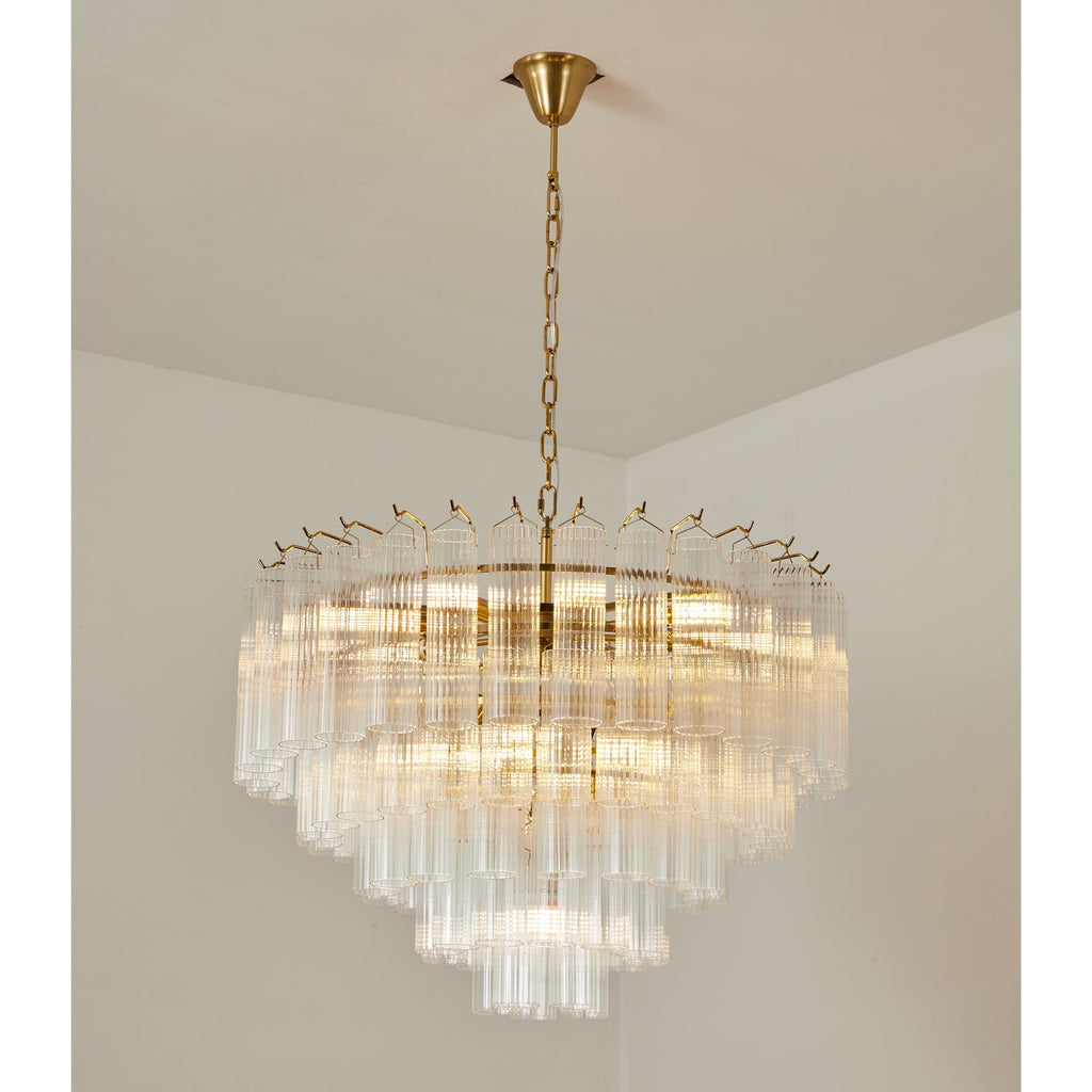 Large lit Clérachic glass chandelier with a gold-toned frame, layers of fluted rods glowing warm in a plain corner.