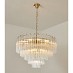 Large lit Clérachic glass chandelier with a gold-toned frame, layers of fluted rods glowing warm in a plain corner.