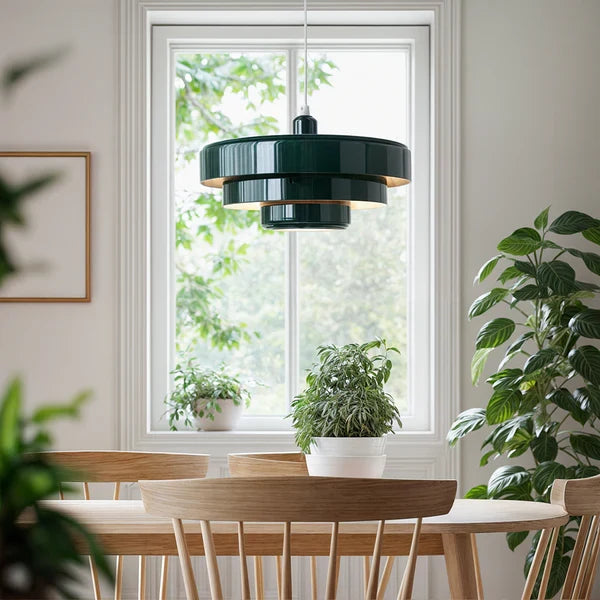 A dark green Clérachic tiered pendant hanging in front of a white-framed window above a wooden table, with leafy plants around it.