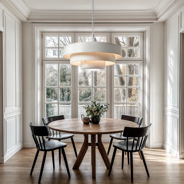 A white Clérachic tiered pendant hanging over a round wooden dining table with black chairs, in a bright room with tall French windows.