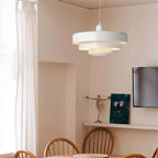 A white Clérachic tiered pendant in the corner of a peach-toned dining room, with wooden chairs, a long curtain and a wall-mounted screen.