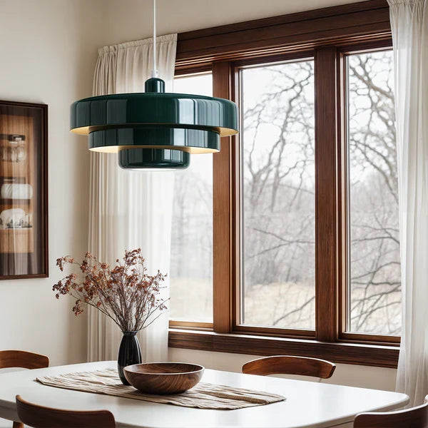 A dark green Clérachic tiered pendant above a white dining table, with a vase of dried stems, a wooden bowl and a wood-framed window.