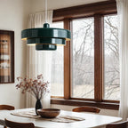 A dark green Clérachic tiered pendant above a white dining table, with a vase of dried stems, a wooden bowl and a wood-framed window.