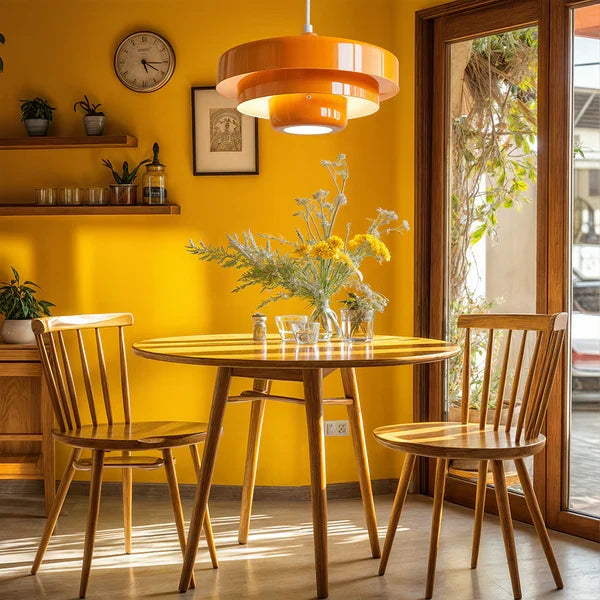 An orange Clérachic tiered pendant in a sunny yellow dining room, above a round wooden table with spindle-back chairs and a wall clock.