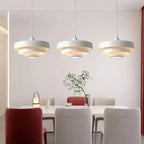 A row of three white Clérachic tiered pendants hanging over a long white dining table with grey and red chairs.