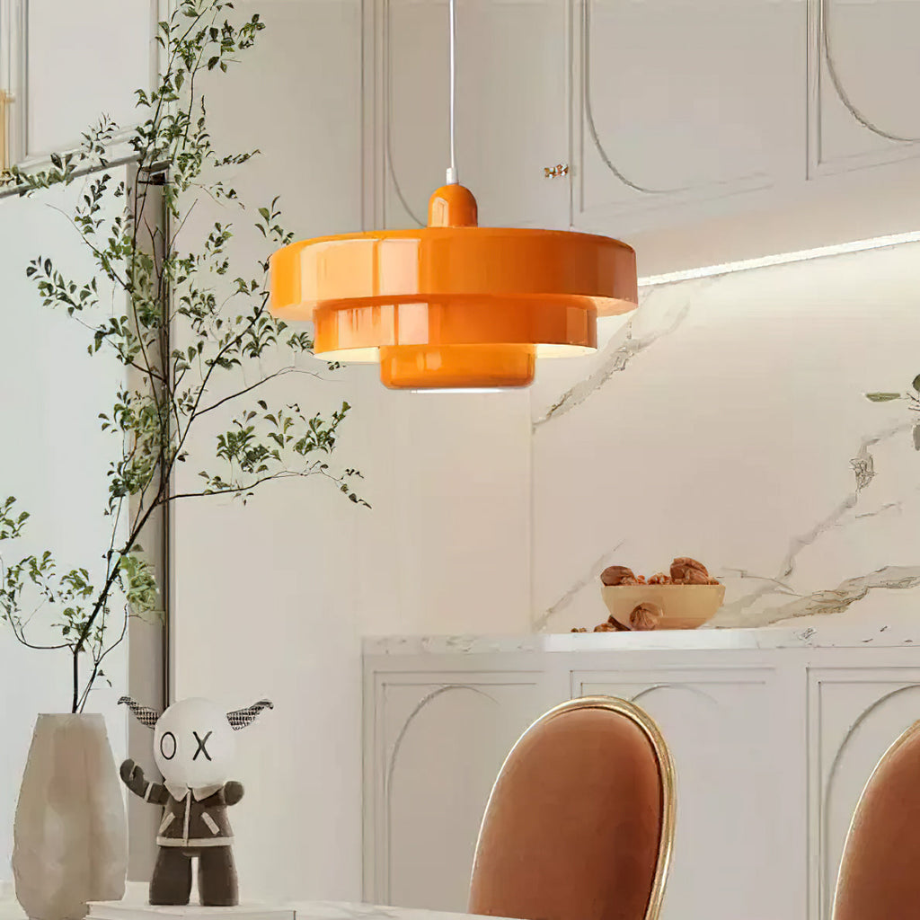 An orange Clérachic tiered pendant on a white cord in a bright kitchen, beside a leafy branch, a marble splashback and a tan chair.