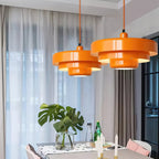 Two orange Clérachic tiered pendants on orange cords in front of sheer curtains, above a dining table with a monstera in a vase.