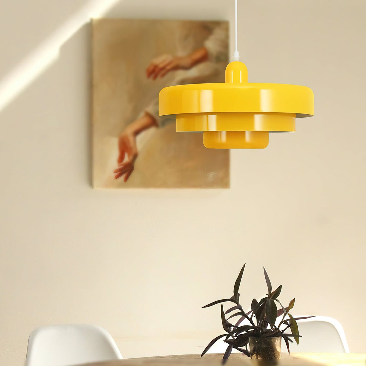 A yellow Clérachic tiered pendant in front of a framed painting of hands, with a plant and white chairs below.