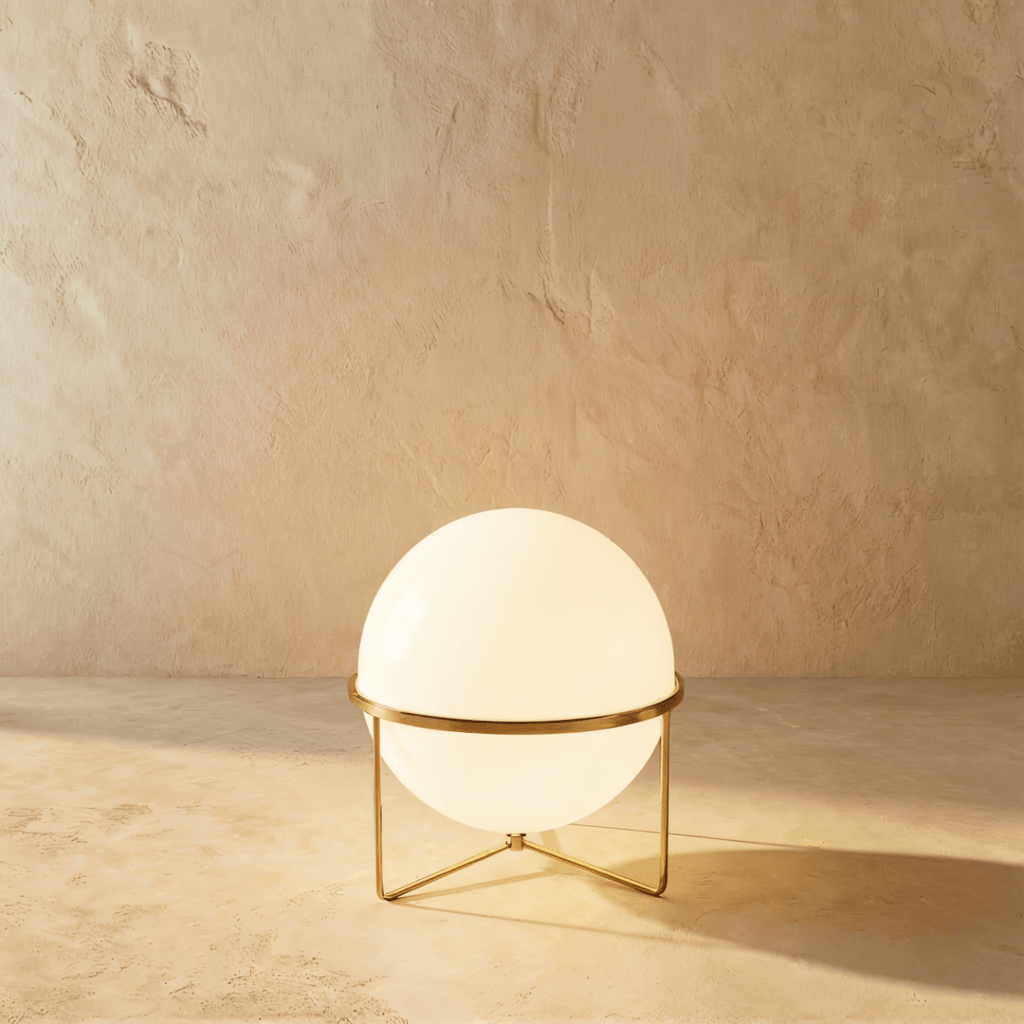 Clérachic single-globe sphere lamp in polished brass, its white globe lit on a three-leg wire foot against a sandy plaster wall.