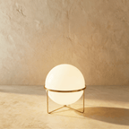 Clérachic single-globe sphere lamp in polished brass, its white globe lit on a three-leg wire foot against a sandy plaster wall.