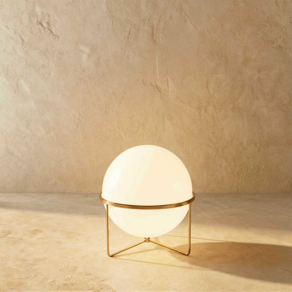 Clérachic single-globe sphere lamp in polished brass, its white globe lit on a three-leg wire foot against a sandy plaster wall.