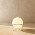 Clérachic single-globe sphere lamp in silver, glowing warm on its wire tripod against the same plaster backdrop.