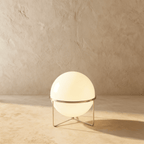 Clérachic single-globe sphere lamp in silver, glowing warm on its wire tripod against the same plaster backdrop.