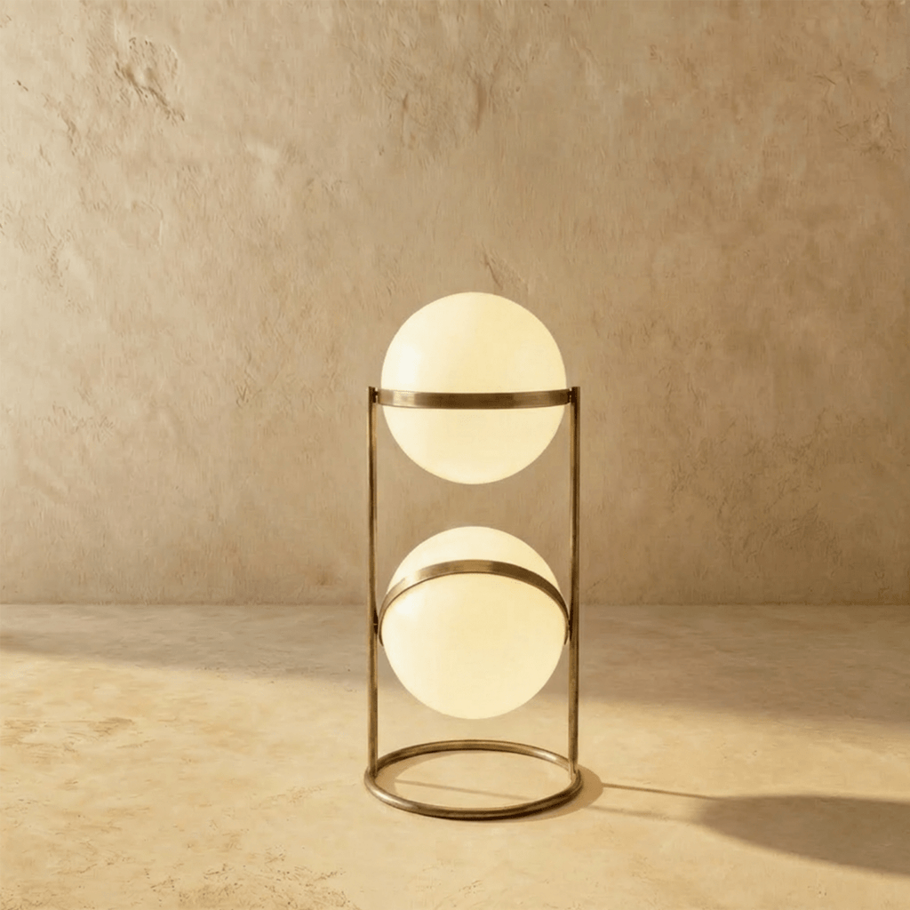 Clérachic two-globe sphere lamp in vintage brass, the short column lit, its darker metal warm against the sandy backdrop.