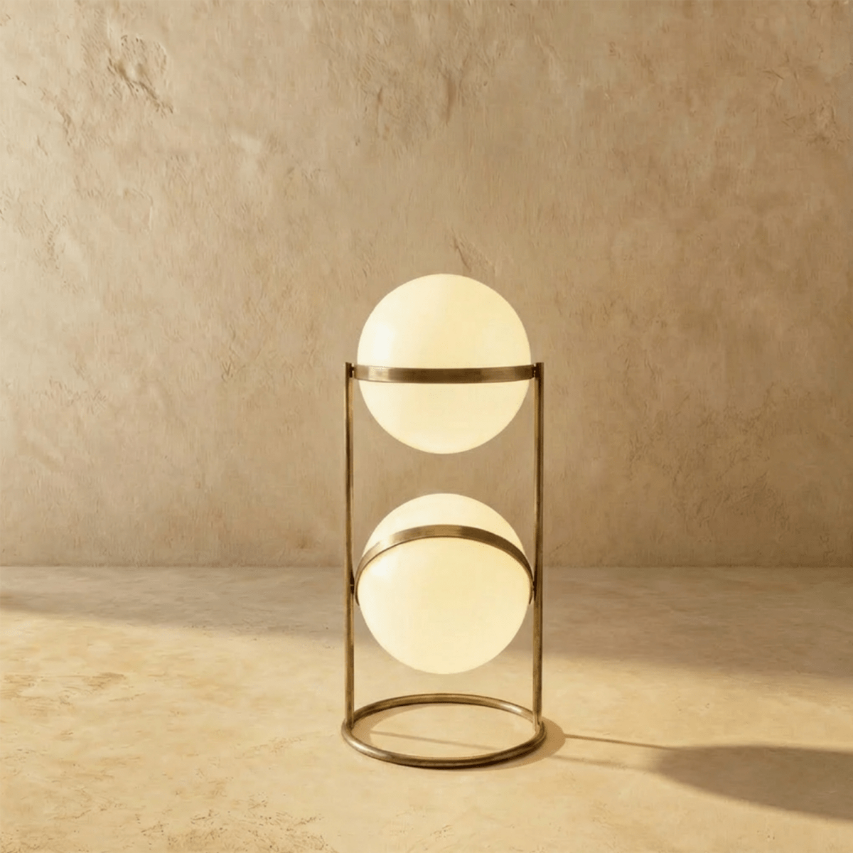Clérachic two-globe sphere lamp in vintage brass, the short column lit, its darker metal warm against the sandy backdrop.