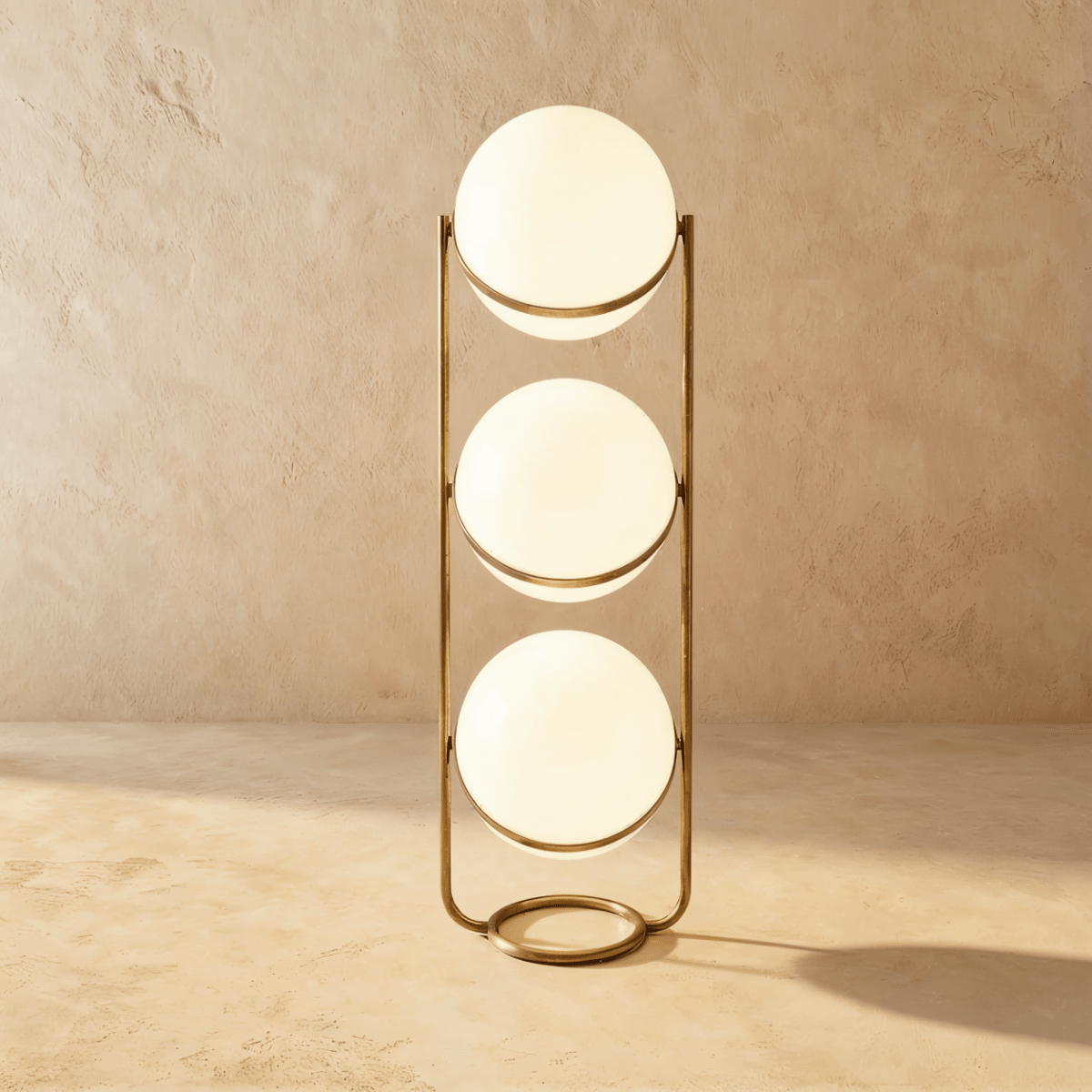 Clérachic three-globe sphere lamp in vintage brass, lit, standing on the sandy plaster studio backdrop.