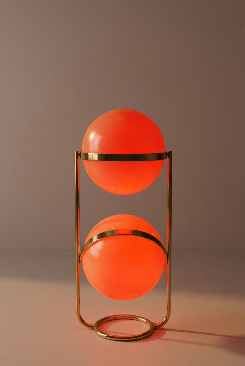 Clérachic two-globe sphere lamp in brass, its globes glowing deep orange against a mauve-grey backdrop.