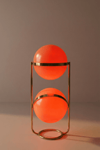 Clérachic two-globe sphere lamp in brass, its globes glowing deep orange against a mauve-grey backdrop.
