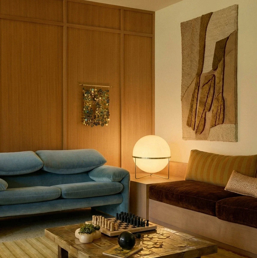 Clérachic single-globe sphere lamp in brass-toned metal, lit on a pale wood plinth in a wood-panelled living room between a blue velvet sofa and a brown velvet daybed.