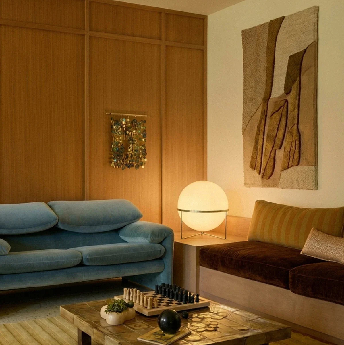Clérachic single-globe sphere lamp in brass-toned metal, lit on a pale wood plinth in a wood-panelled living room between a blue velvet sofa and a brown velvet daybed.