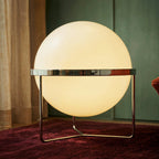 Clérachic single-globe sphere lamp standing on a deep red rug, its mirror-polished band reflecting the green panelled wall behind it.