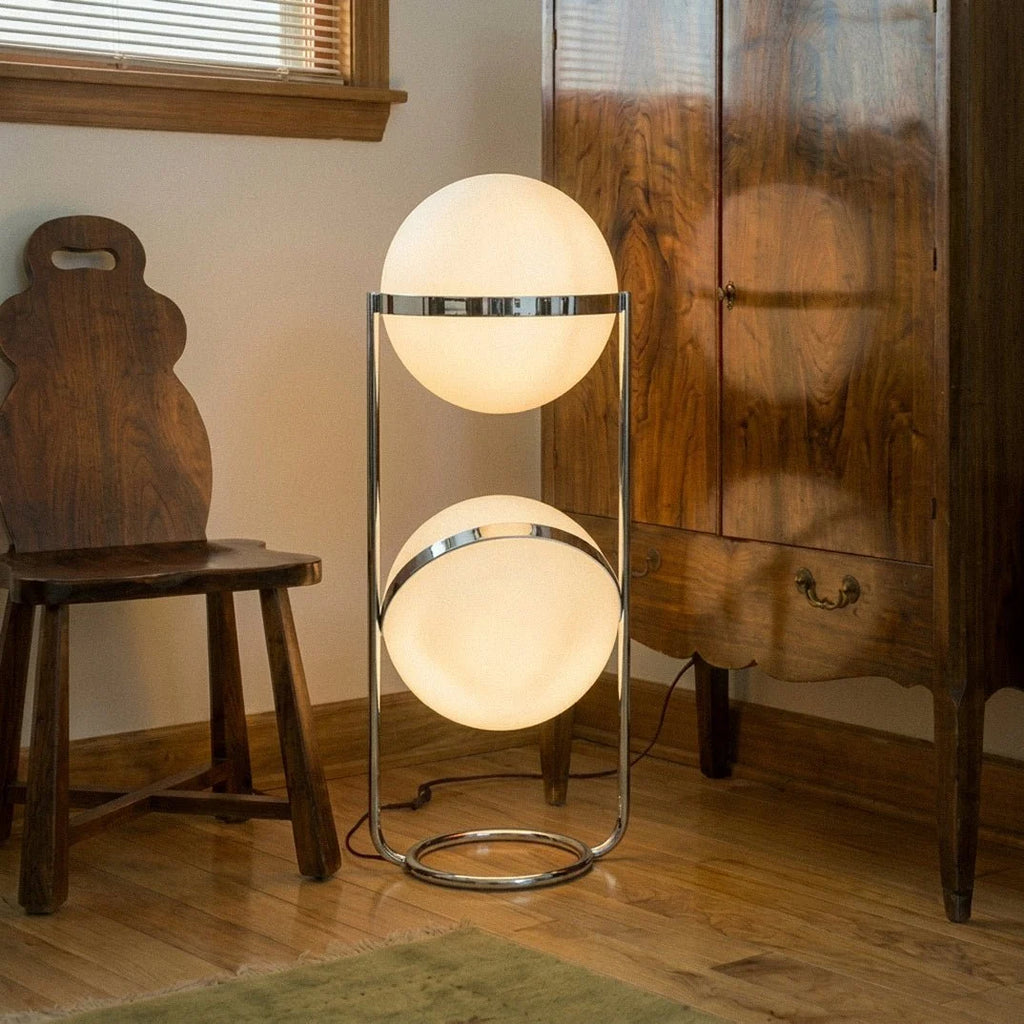 Clérachic two-globe sphere lamp in mirror-polished silver, lit on a wood floor by a walnut cabinet, its cord running right and behind the furniture.